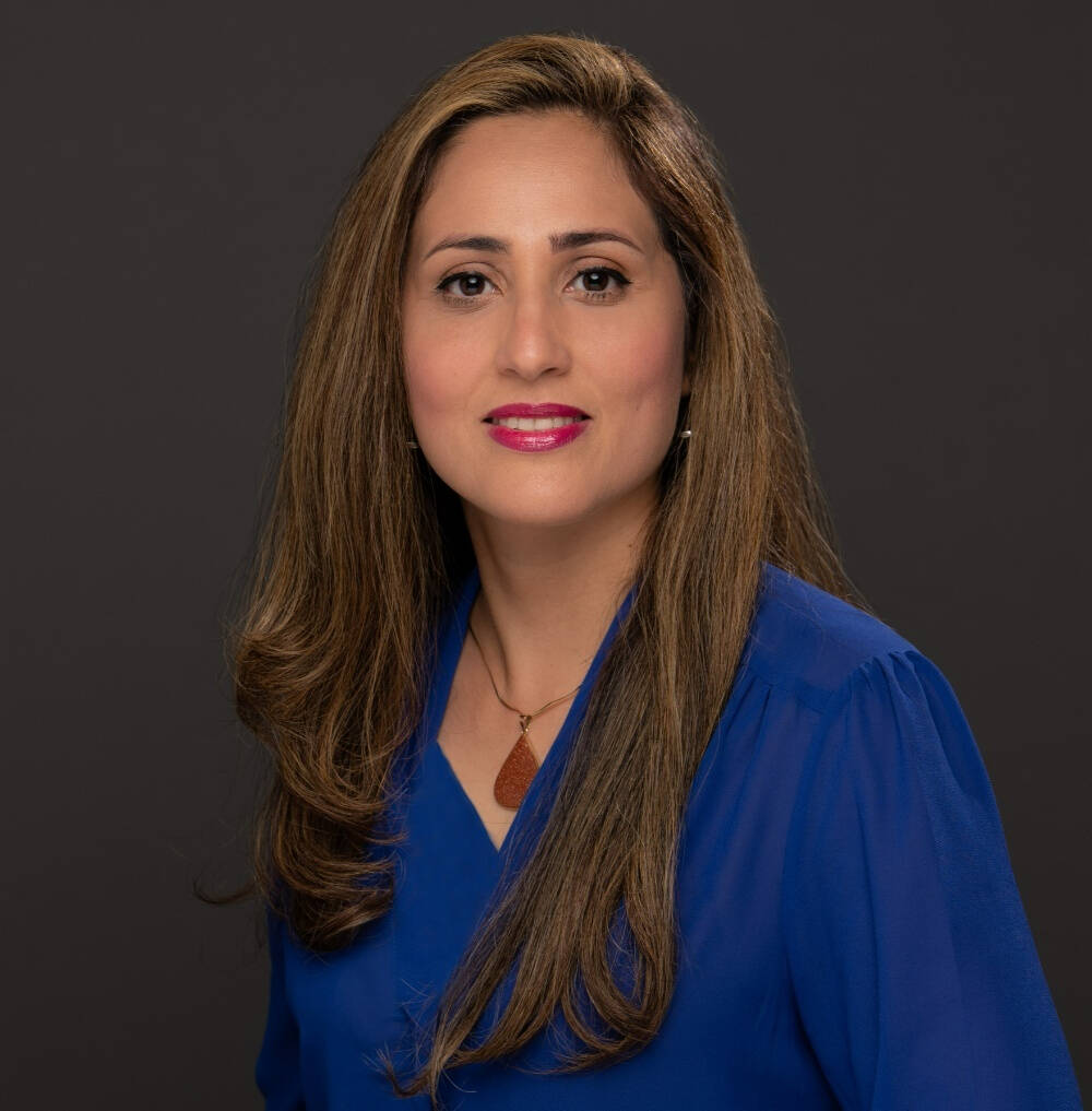 Portrait of a woman with long hair and a blue blouse, looking directly at the camera with a neutral expression. The dark gray background emphasizes her face and attire.